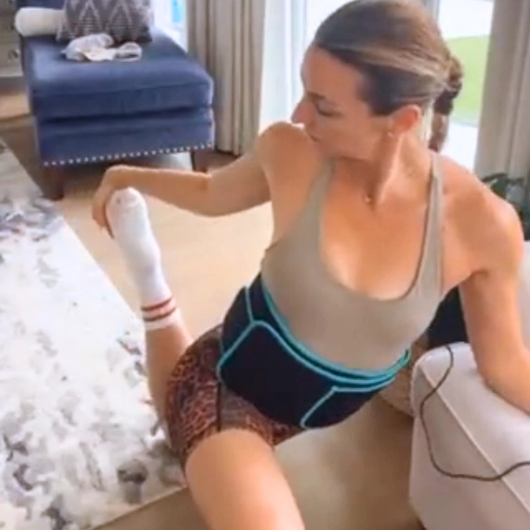 Térdelő, a lábát nújtó nő derékon viseli a vörös fényterápiás szalagot, a nappalijában edzés közben.