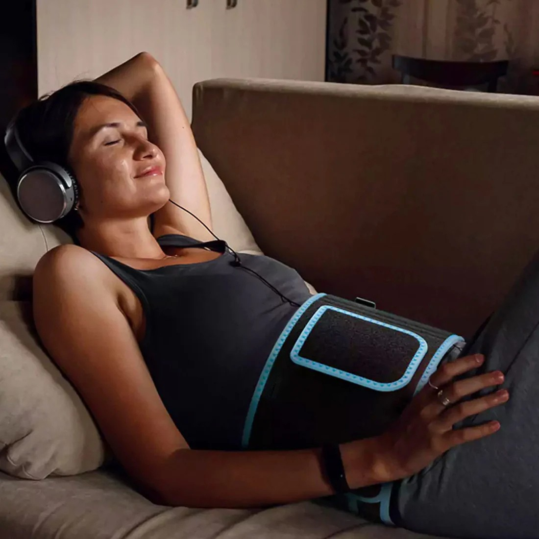 Nő pihen a kanapén, hasán a fényterápiás szalag; otthoni, relaxáló használat, napi rutin.